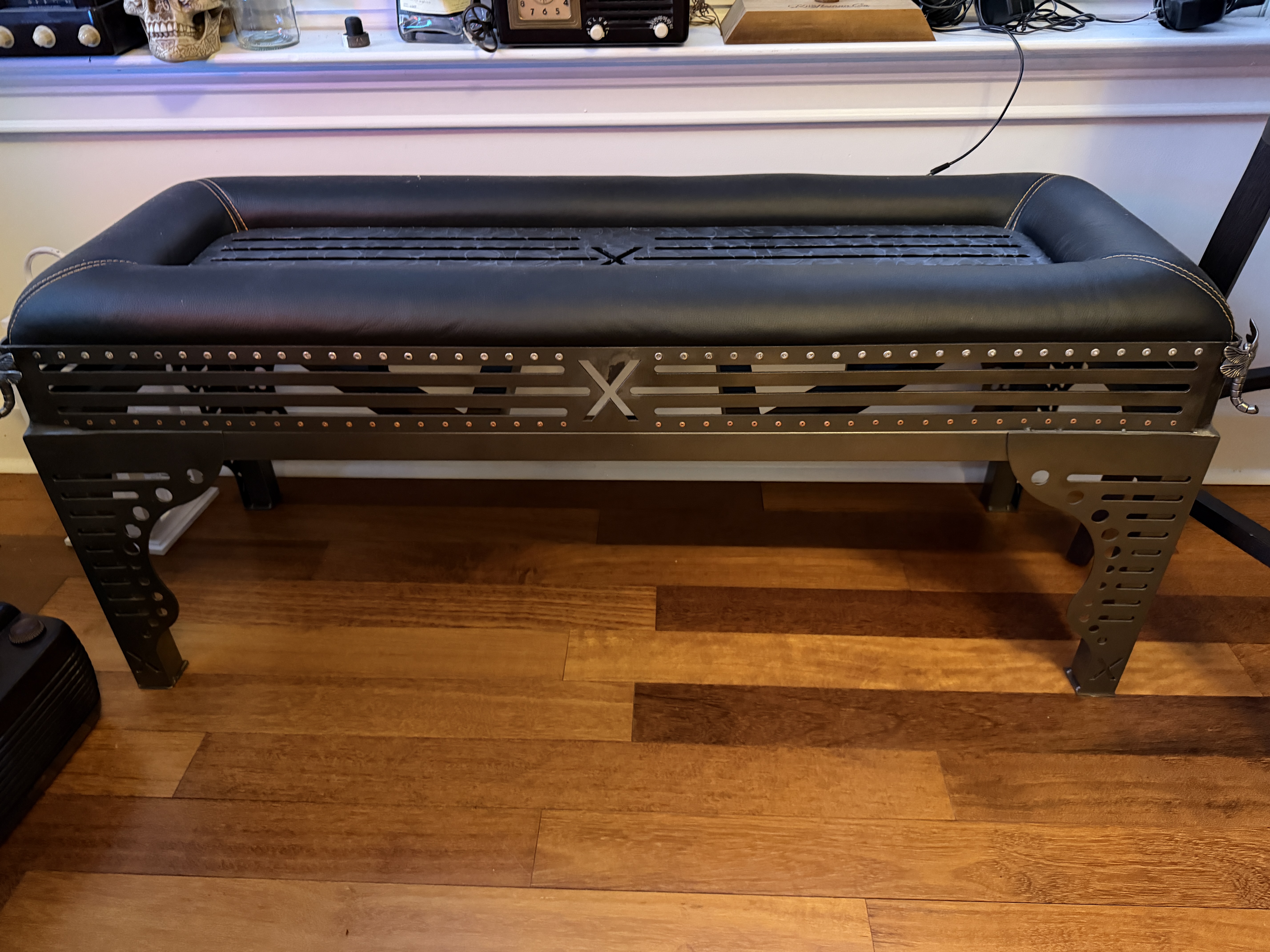 A custom bench with a black leather-topped seat and a hand-cut riveted steel frame, signed with Jon's X mark on the front panel.